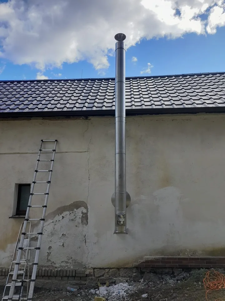 Nerezový komín vedený po stěně staršího hospodářského stavení nad hřeben střechy
