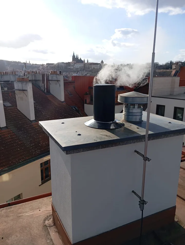 Kouřící komínové vyústění na střeše pražského domu, v&nbsp;pozadí panorama Hradčan