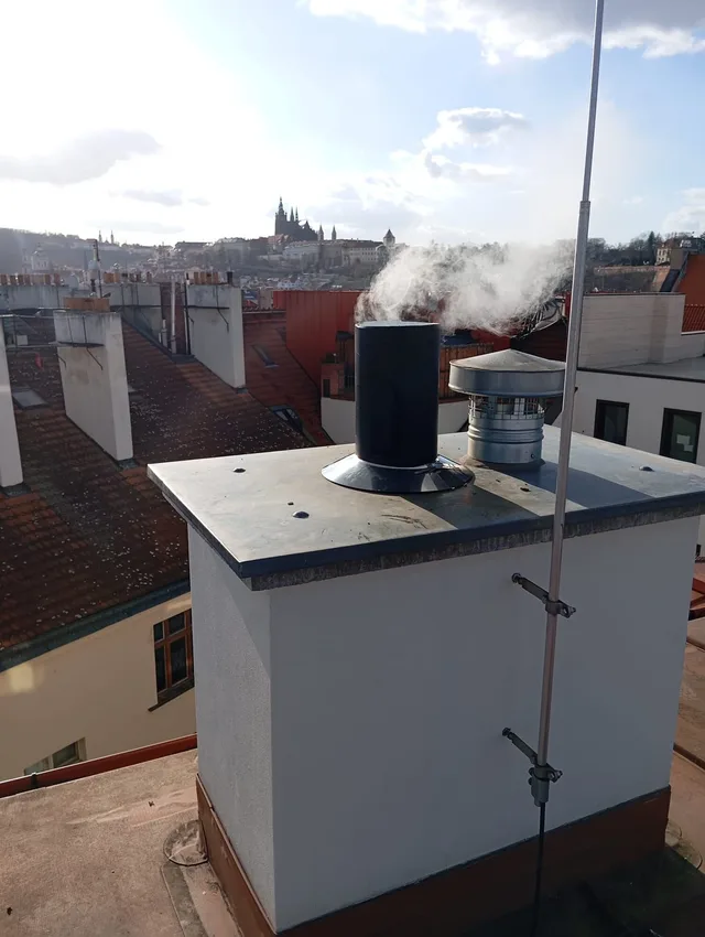 Kouřící komínové vyústění na střeše pražského domu, v&nbsp;pozadí panorama Hradčan