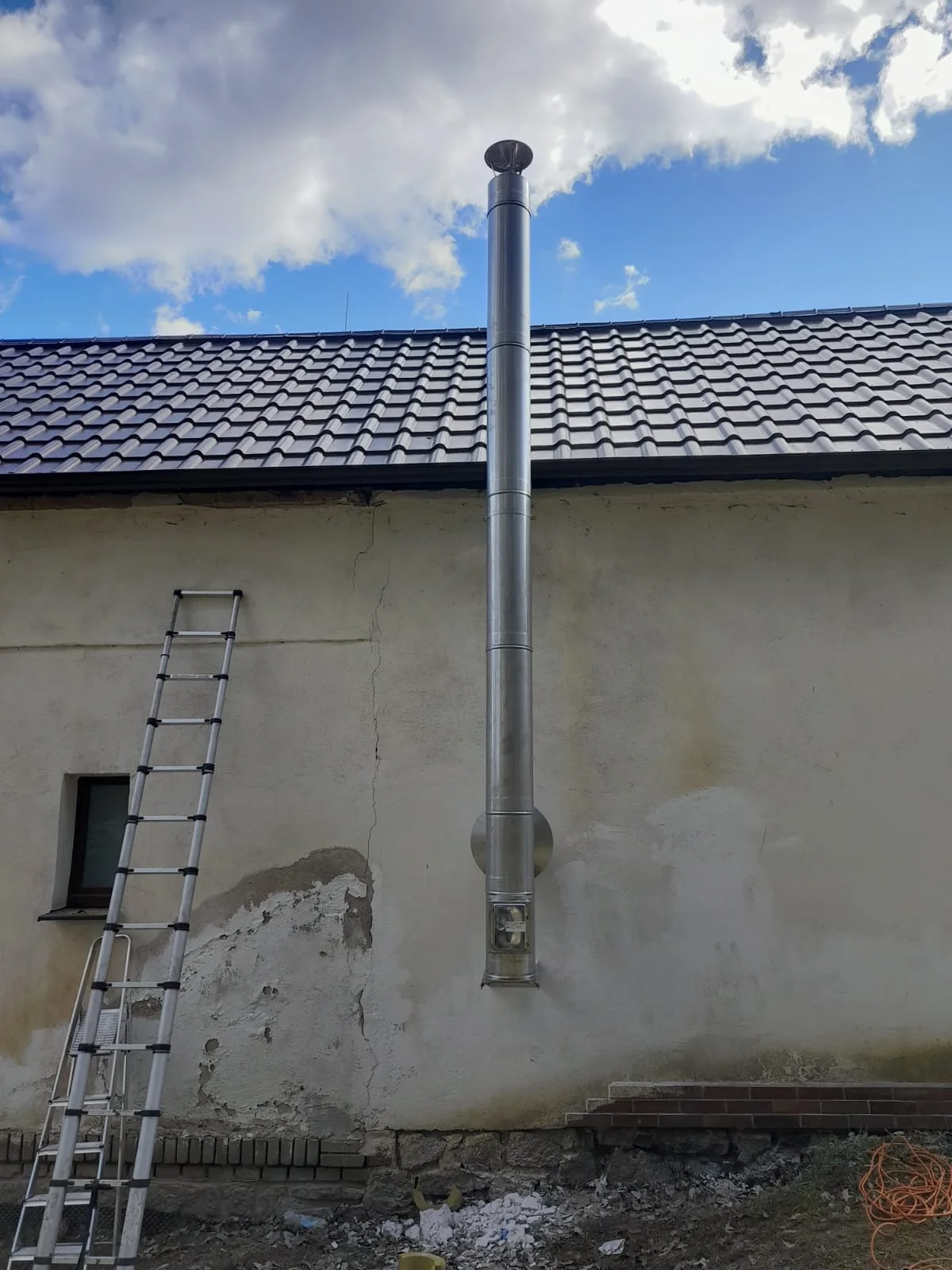 Žebřík opřený o stěnu domu vedle nerezového komína vedeného po fasádě
